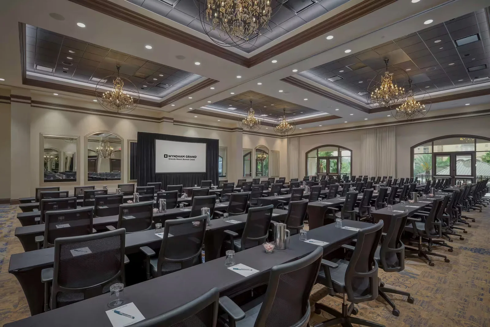 Formal conference room with rows of tables and chairs facing a presentation screen, ornate chandeliers, and large windows.
