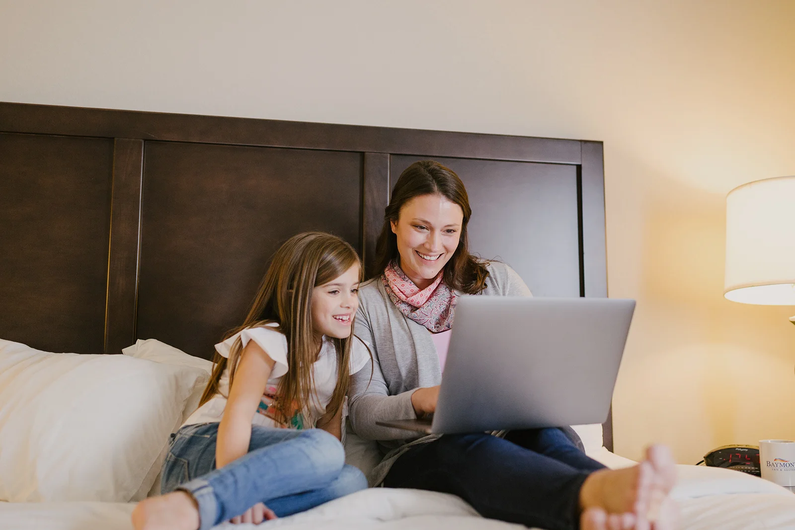 Woman and young girl smiling while using a laptop together on a bed.