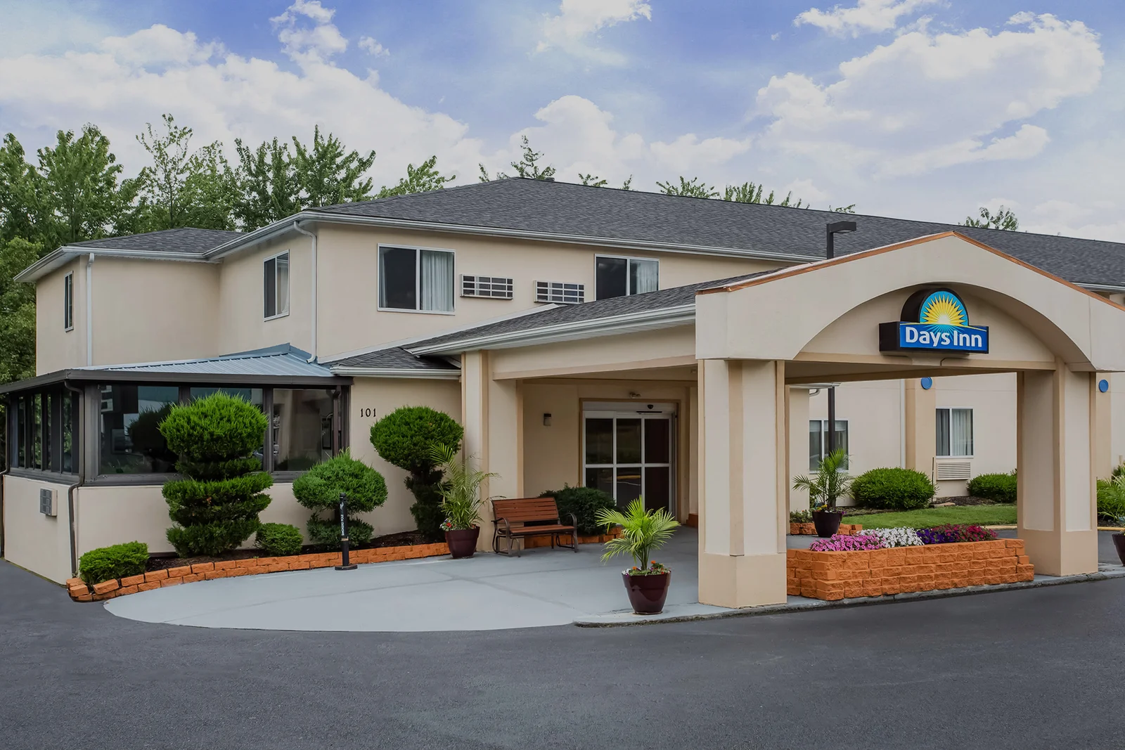 Exterior of a two-story Days Inn hotel with a covered entrance, landscaped shrubs and flower beds and a blue sky with clouds 