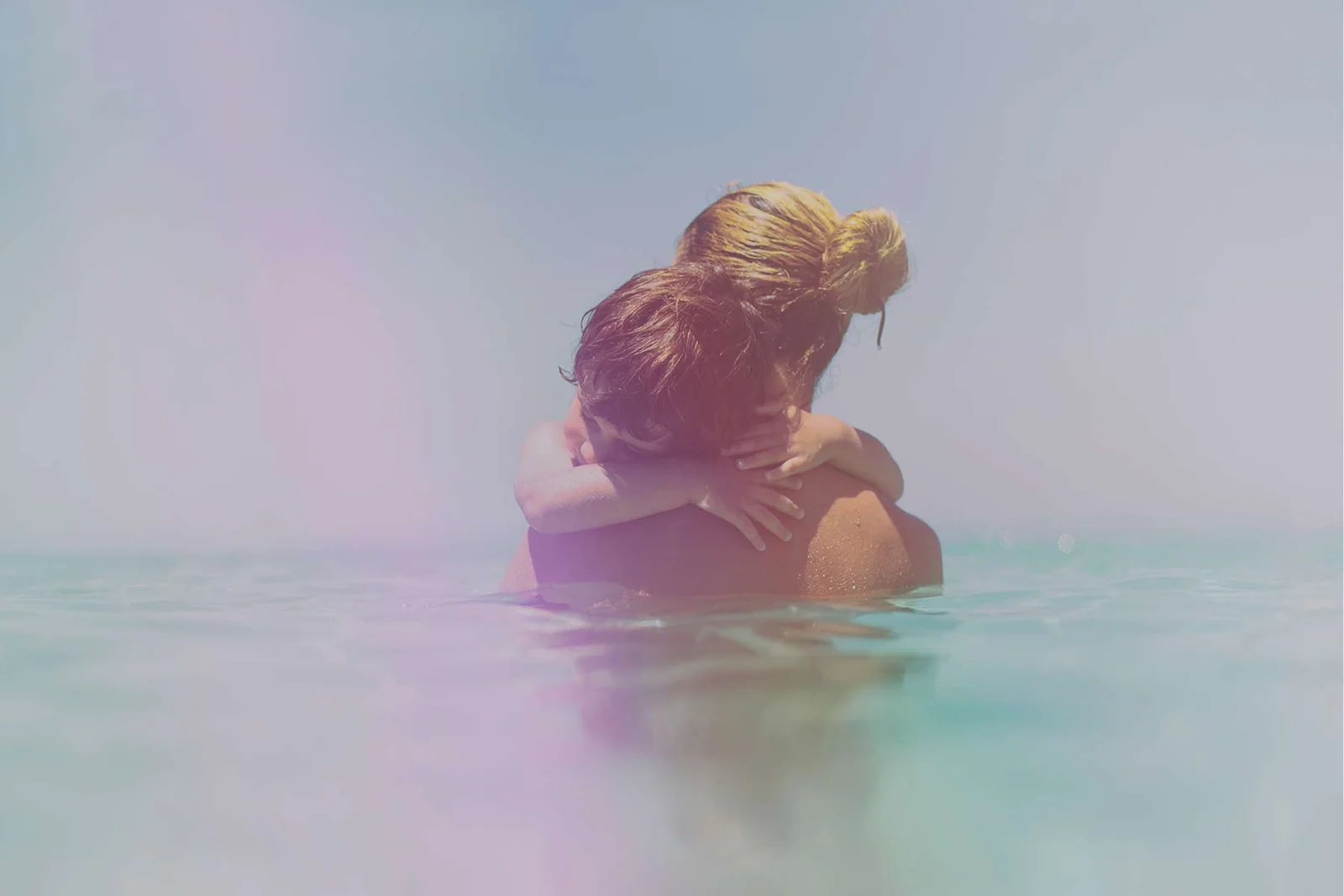 Two people embrace in calm, pastel-colored water, with one person’s reddish-brown hair tied in a loose bun.