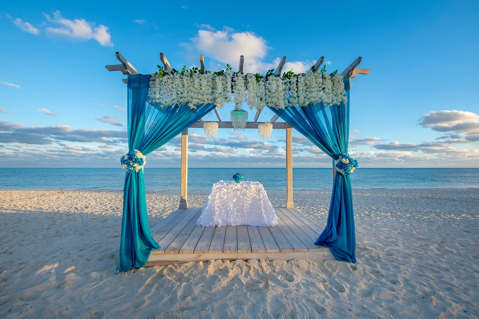 A teal-draped decorative canopy with foliage and a white-covered table stands on a sandy beach beside the ocean.