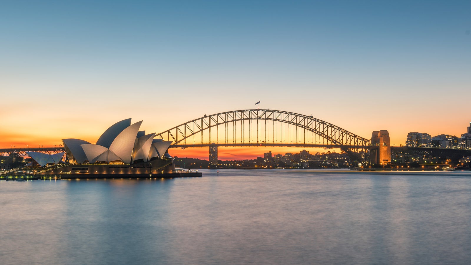 Sydney Opera House and Harbour Bridge silhouetted against a colorful sunset over calm water.