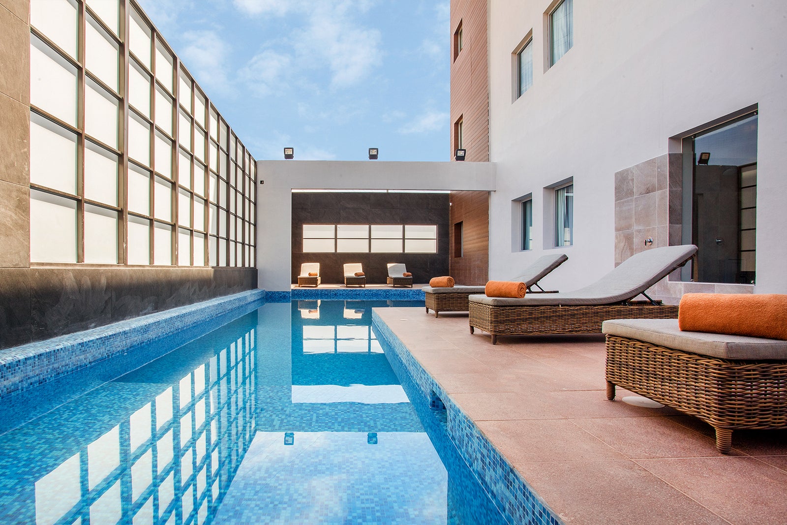 Modern indoor courtyard pool with blue mosaic tile, reflective water, and wicker lounge chairs beside large grid windows.