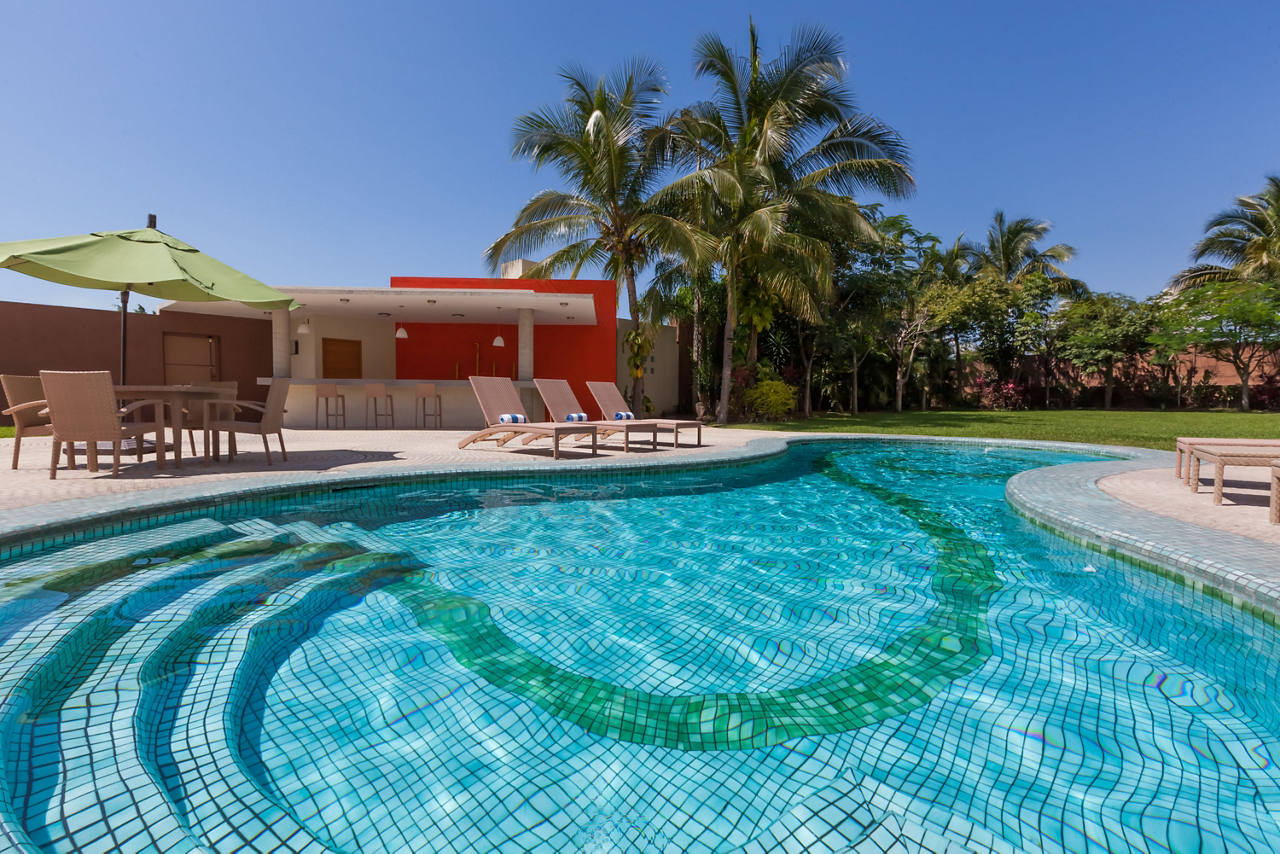 Sunlit tiled swimming pool beside a modern outdoor bar, lounge chairs, patio furniture, and palm trees.
