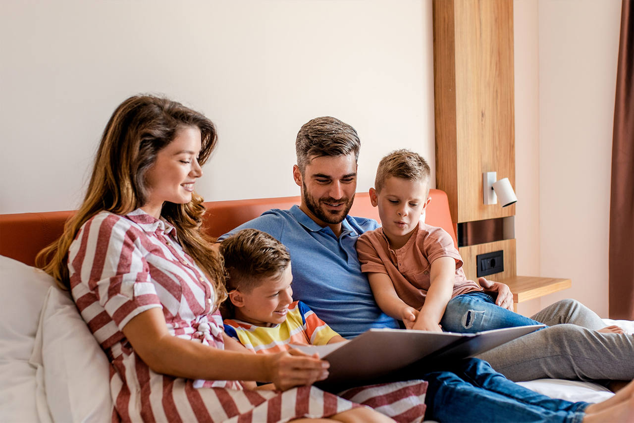 A family of mom, dad, two boys, huddled over a large children's book on a soft, pillowed bed.