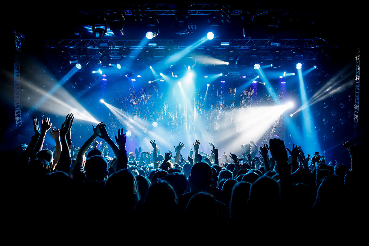 A packed concert with beams of light shining across the stage of a dark venue.
