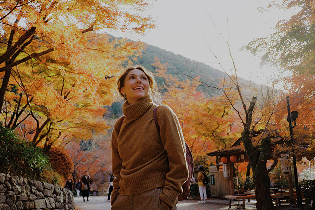 Smiling person in a brown sweater on a path surrounded by brilliant autumn foliage, with a forested mountain in the background.